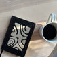 White mug with black coffee on a wooden surface next to a black notebook with white patterns