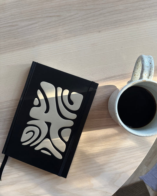 White mug with black coffee on a wooden surface next to a black notebook with white patterns
